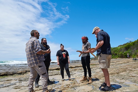 Five First Nations People stand in a semi circle on the tidal rocks on Gadubanud Country. Two men hold boomerangs.  There is one woman in the centre, they are all engaged in a discussion.  It is a sunny day at low tide.